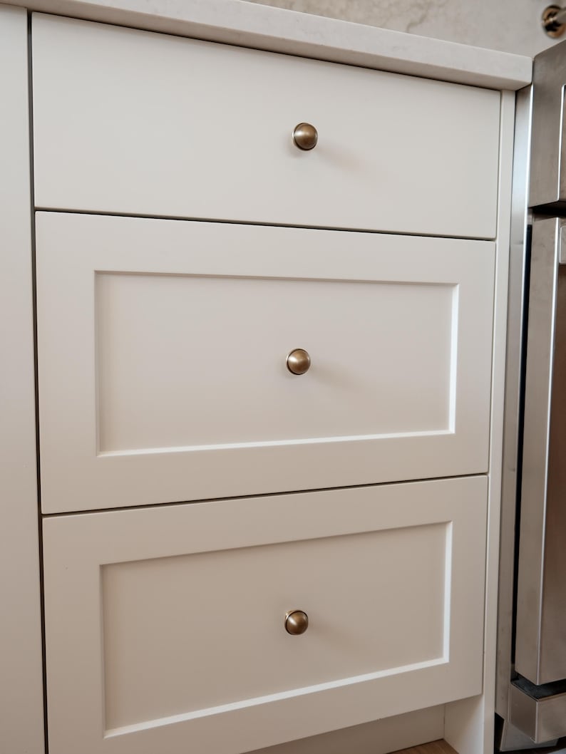 May include: Close-up of three off-white kitchen cabinet drawers with brass-coloured knobs. The drawers are rectangular with a recessed panel design. The top drawer is plain, while the bottom two have a framed appearance. The cabinets are part of a larger kitchen setup.