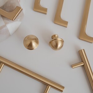 May include: A selection of gold-coloured cabinet hardware, including handles and knobs, arranged on a white surface. The hardware has a matte finish and a modern design. A decorative marble bowl with a checkered pattern is also visible.