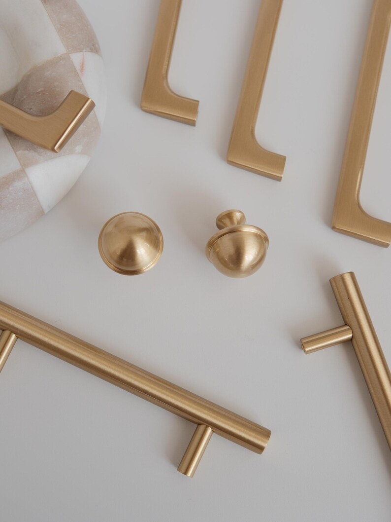 May include: A selection of gold-coloured cabinet hardware, including handles and knobs, arranged on a white surface. The hardware has a matte finish and a modern design. A decorative marble bowl with a checkered pattern is also visible.