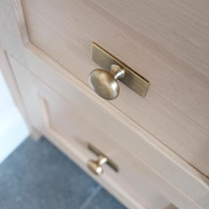 May include: Close-up of a light-coloured wooden cabinet with two drawers. Each drawer has a brass-coloured handle. The top handle is a knob attached to a rectangular backplate. The bottom handle is a smaller, similar design.