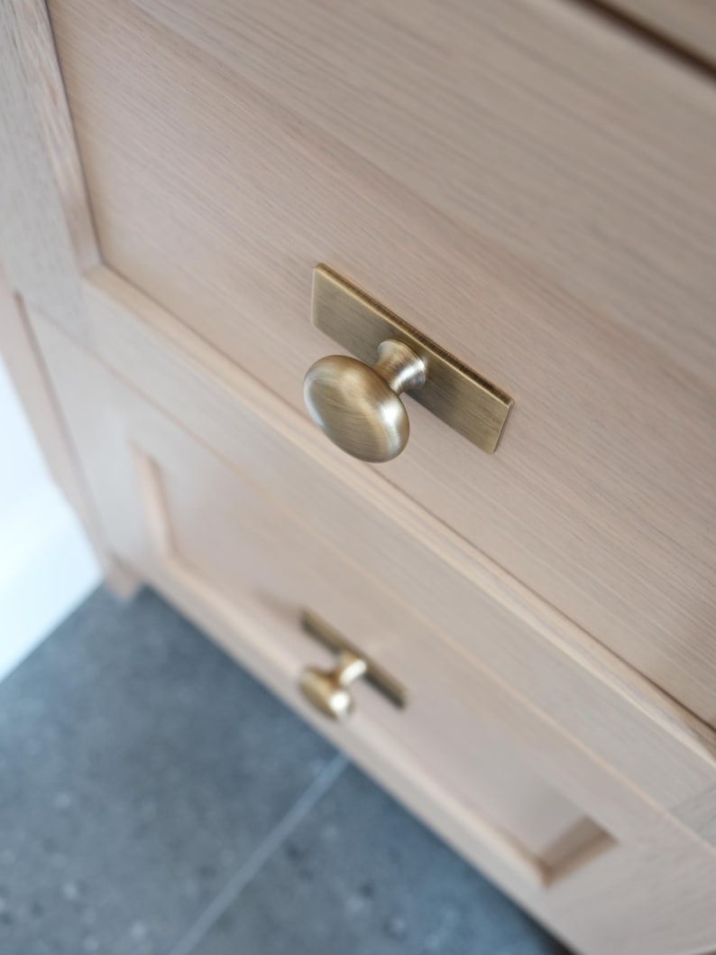 May include: Close-up of a light-coloured wooden cabinet with two drawers. Each drawer has a brass-coloured handle. The top handle is a knob attached to a rectangular backplate. The bottom handle is a smaller, similar design.