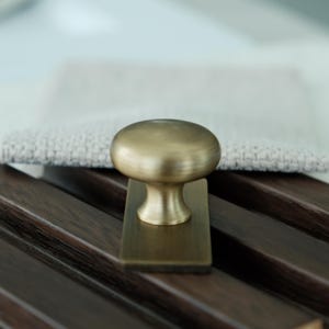 May include: A brass cabinet knob with a rounded top and rectangular base. The knob is set against a dark wood surface with a light-coloured fabric in the background. The hardware has a vintage aesthetic.