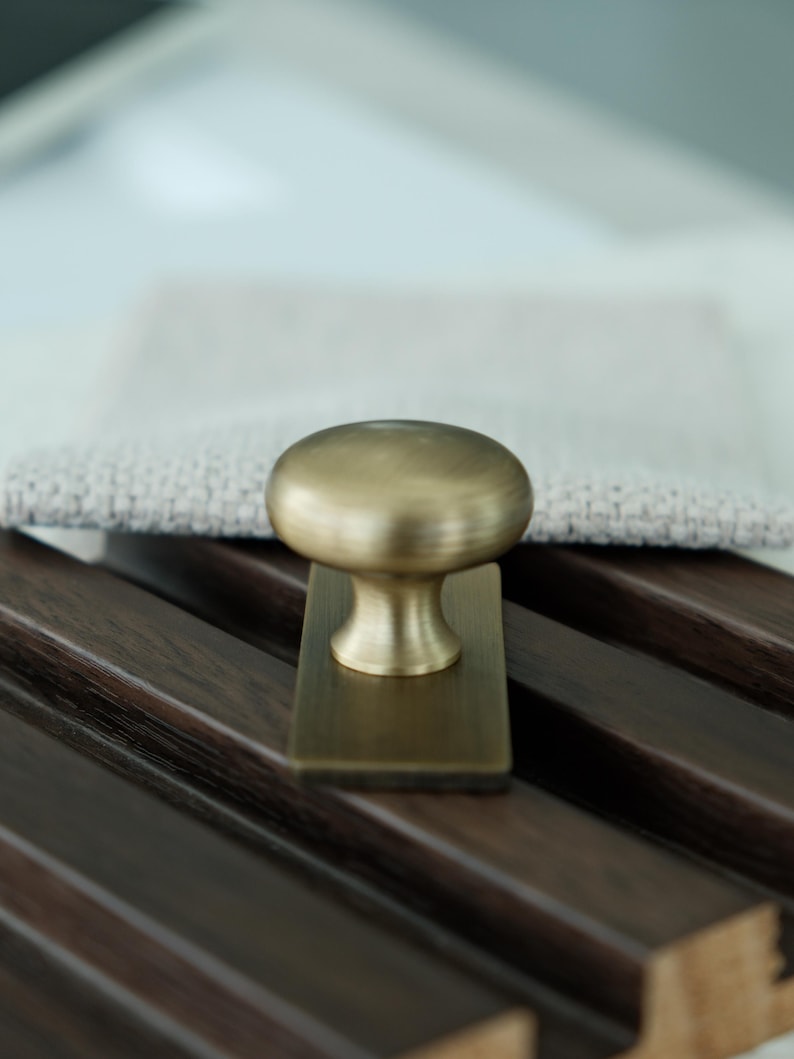 May include: A brass cabinet knob with a rounded top and rectangular base. The knob is set against a dark wood surface with a light-coloured fabric in the background. The hardware has a vintage aesthetic.