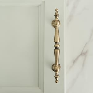 Peut inclure: Une poignée de meuble en laiton antique. La poignée est orientée verticalement et présente un design décoratif et orné. La poignée est montée sur une porte d'armoire blanche et un mur en marbre blanc.