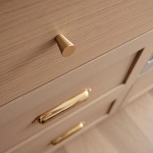 May include: Close-up of light wood cabinetry with gold-toned hardware. The top drawer features a conical knob, while the lower drawers have elongated, curved pulls. The hardware contrasts with the natural wood grain.