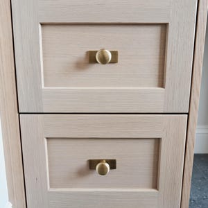 May include: Close-up of a light-coloured wooden cabinet with two drawers. Each drawer has a rectangular brass pull with a round knob. The cabinet has a simple, modern design, with visible wood grain.