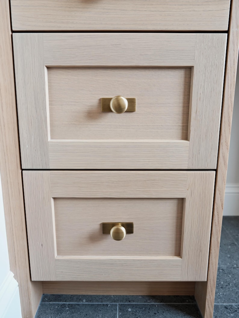 May include: Close-up of a light-coloured wooden cabinet with two drawers. Each drawer has a rectangular brass pull with a round knob. The cabinet has a simple, modern design, with visible wood grain.