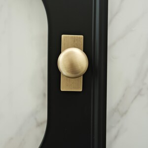 May include: Close-up of a black door or cabinet with a brass-coloured knob. The knob is round and sits on a rectangular backplate. The background is a white marble-like surface.