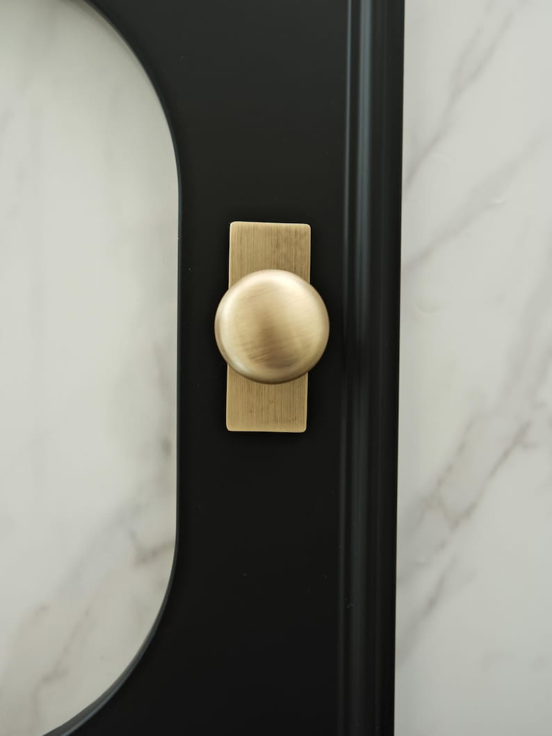 May include: Close-up of a black door or cabinet with a brass-coloured knob. The knob is round and sits on a rectangular backplate. The background is a white marble-like surface.