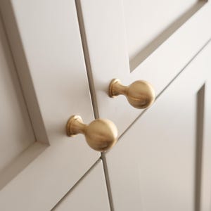 May include: Close-up of two gold-colored cabinet knobs with a spherical design. The knobs are mounted on a light gray cabinet door with a recessed panel design. The hardware adds a touch of elegance to the cabinetry.