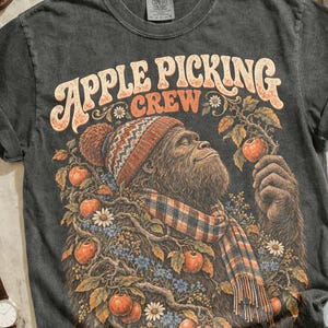May include: A charcoal gray t-shirt featuring a graphic of a creature wearing a patterned beanie and scarf, surrounded by apples and the text Apple Picking Crew. The shirt is displayed with brown leather boots, a watch, camera, and sunglasses.