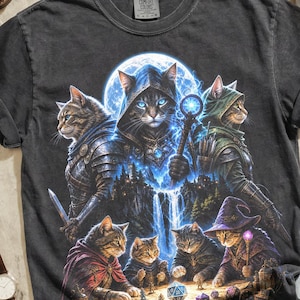 May include: Graphite-colored t-shirt featuring a fantasy cat design. The graphic depicts cats in wizard and knight attire, with a full moon backdrop. The shirt is styled with brown leather boots, a watch, sunglasses, and a camera.
