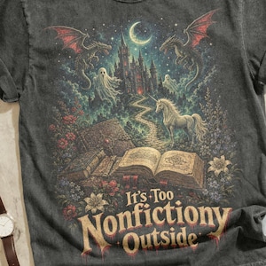 May include: A charcoal gray t-shirt featuring a fantasy graphic of a castle, dragons, a unicorn, and an open book with the text "It's Too Nonfictiony Outside." The shirt is surrounded by brown leather boots, a camera, a watch, sunglasses, and a chain.