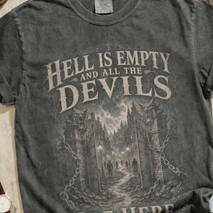 May include: A charcoal grey short-sleeve t-shirt featuring a graphic of a gothic gate with the text Hell is empty and all the devils are here. The shirt is surrounded by brown leather boots, a silver chain, a camera, a watch, jeans, and sunglasses.