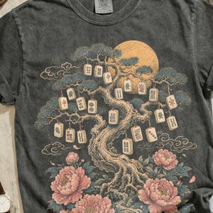 May include: A charcoal gray t-shirt featuring a graphic of a tree with mahjong tiles hanging from its branches, surrounded by pink peonies and a yellow moon. The shirt is displayed with brown leather boots, a silver chain, a camera, a watch, sunglasses, and light blue jeans.