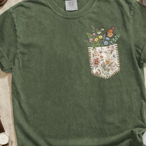 May include: A moss green t-shirt with a pocket featuring a floral and butterfly design. The image includes brown leather boots, a watch, sunglasses, a camera, and a necklace. The color of the shirt is labeled "Color Moss".