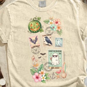 May include: Ivory-colored t-shirt featuring a collage of Halloween-themed postage stamps with a pumpkin, bat, raven, and ghost. The shirt is surrounded by accessories like boots, a watch, sunglasses, and a camera.