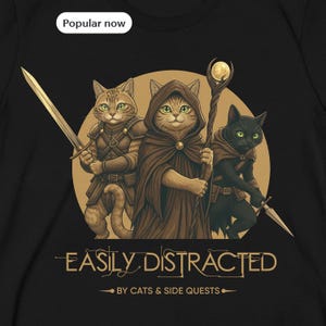 Op de afbeelding: Zwart t-shirt met een fantasie-thema afbeelding van drie katten. Eén kat houdt een zwaard vast, een andere een staf en de derde een dolk. De tekst "EASILY DISTRACTED" en "BY CATS & SIDE QUESTS" staat eronder.