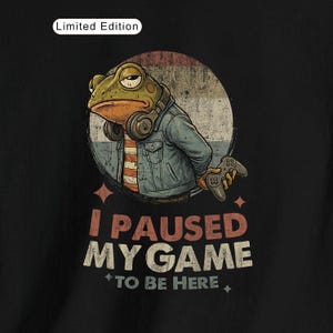 Peut inclure: T-shirt noir avec une grenouille de dessin animé portant une veste en jean et un casque, tenant une manette de jeu. Le texte "I PAUSED MY GAME TO BE HERE" est affiché en dessous. Les mots "Limited Edition" sont en haut.