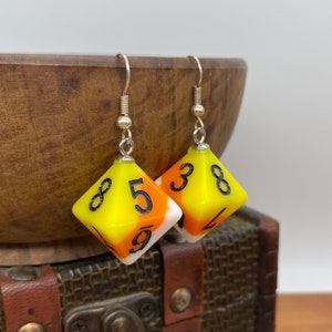 May include: Earrings with yellow, orange, and white dice-shaped charms, each with black numbers. The charms hang from silver-coloured hooks. The earrings are set against a wooden bowl and a vintage-style chest, creating a playful aesthetic.