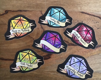 May include: A collection of six colorful D20 dice stickers with black outlines. Each die features a different color scheme and a banner with phrases like "Lucky Me" and "Critical Success". The dice are set against a wooden background.