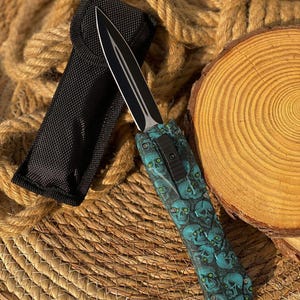 Può includere: Un pugnale a doppio filo con lama nera dotata di scanalatura centrale e impugnatura decorata con un motivo di teschi color verde acqua con occhi verde brillante. Il coltello include un fodero in nylon nero con chiusura a patta e bottone.