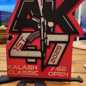 Puede incluir: Un letrero de madera rojo y negro con el texto "AK 47 1st 2025" y "Kalash Classic 7.62 Open". El letrero presenta un gráfico de un rifle. El letrero está sobre una superficie de madera.