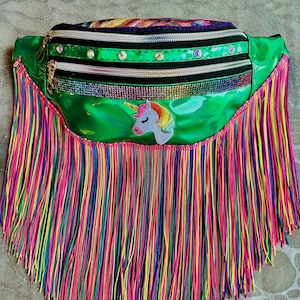 Puede incluir: Un riñonera verde vibrante con flecos de arcoíris. La riñonera presenta un parche de unicornio, una cremallera negra y pedrería decorativa. La parte superior de la riñonera tiene un diseño a rayas colorido.