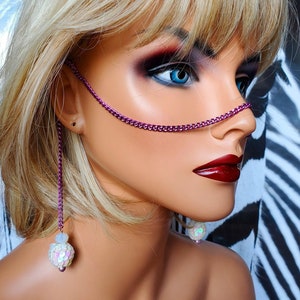 May include: A mannequin head with a purple chain nose ring and dangling earrings. The earrings have a white and iridescent sequin design. The mannequin has blonde hair, blue eyes, and dark red lipstick. The background is a black and white zebra print.