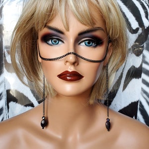 May include: A mannequin head with blonde hair and dramatic eye makeup. It is wearing a black chain mask and long chain earrings with black beads. The background features a zebra print.