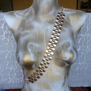 May include: A gold-toned necklace with a rectangular link design is draped across a mannequin. The necklace is made of a series of connected, rectangular links. The mannequin is painted with gold and white tones.