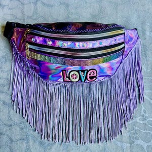 Peut inclure: Un sac banane holographique avec une frange violette. Le sac comporte plusieurs compartiments zippés avec des accents irisés et des strass. Le mot "LOVE" est brodé sur le devant en lettres colorées.