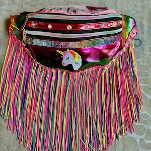 May include: A colorful fanny pack with a rainbow fringe. The bag has a holographic finish and features a unicorn patch. The zippers are black and the bag is decorated with rhinestones. The fringe is made of multiple colors.