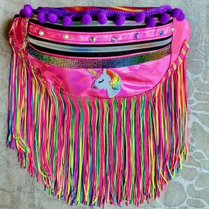 May include: A vibrant pink fanny pack with a rainbow fringe and a unicorn patch. The bag features a black zipper, decorative rhinestones, and purple pom-poms along the top edge. The fringe is a mix of pink, yellow, green, and blue.