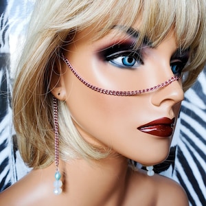 May include: A mannequin head with a pink chain nose and ear jewelry. The chain nose piece connects to a chain that dangles from the ear. The earrings have a chain with a blue and white bead. The mannequin has blonde hair, blue eyes, and dark red lipstick.