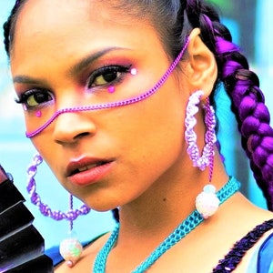 May include: A woman with purple braided hair and matching makeup, including a chain across her face and hoop earrings. The earrings and necklace are a light purple and blue. The woman is wearing a blue top with sequins.