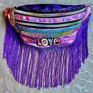 May include: A colourful fanny pack with a holographic finish, decorated with purple fringe. The bag has multiple zipped compartments, embellished with rhinestones and the word "LOVE".