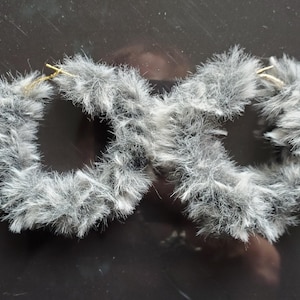 May include: A pair of fluffy gray hoop earrings. The earrings are made of soft, textured material, with a gold-colored hook. The earrings are designed in a circular shape, with a fuzzy, fur-like appearance. The earrings are a fashion accessory.