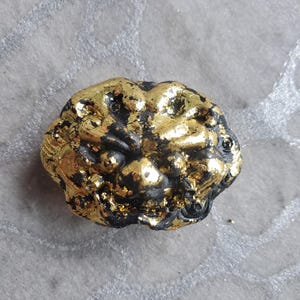 May include: A black and gold amulet with a detailed, textured design. The amulet is shaped like a face with a wide mouth and large eyes.