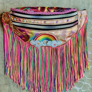 May include: A colorful fanny pack with a rainbow and cloud patch. The bag features a holographic metallic finish, multiple zippered compartments, and a fringe of neon-colored strands. The top edge has a pink trim.