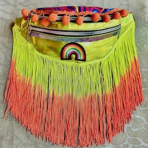 May include: A neon yellow fanny pack with a rainbow patch and a zipper closure. The bag is adorned with orange fringe and pom-poms. The top of the bag has a colorful, abstract pattern.