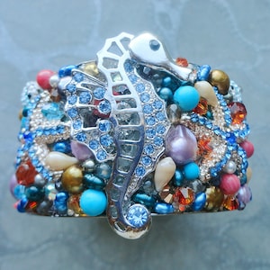 Può includere: Un bracciale a polsino largo con un motivo a cavalluccio marino argentato. Il bracciale è adornato con una varietà di perline e pietre preziose colorate, tra cui accenti blu, turchesi, arancioni e perlati. Il design è ornato e accattivante.