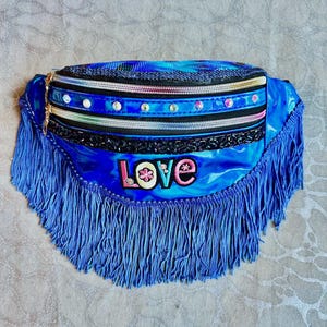 May include: A vibrant blue fanny pack with a shiny, textured finish. The bag features multiple decorative zippers, sparkling embellishments, and a fringe trim. The word "LOVE" is embroidered on the front. A gold-toned zipper closure is visible.