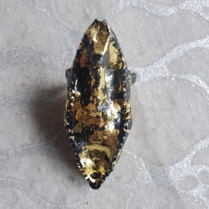 以下が含まれることがあります： 黒と金のリング、テクスチャ加工された表面。 リングは細長く、先端が尖っており、金箔で覆われています。 リングはおそらく金属製で、暗くマットな仕上がりです。