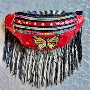 Könnte beinhalten: Rote Bauchtasche mit Schmetterlingsaufnäher, schwarzem Fransenbesatz und mehreren Reißverschlussfächern. Die Bauchtasche hat eine glänzende, reflektierende Oberfläche und ist mit Strasssteinen und einem Regenbogen-Reißverschluss verziert.