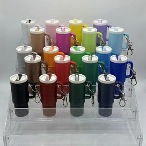 May include: A display of colorful, insulated travel mugs with handles and attached keychains. The mugs come in various colors, including black, red, green, and blue, and are arranged on a clear, tiered display stand.