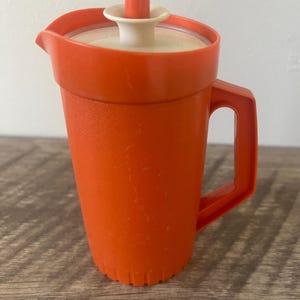 Può includere: Una caraffa in plastica arancione brillante con esterno testurizzato, una robusta maniglia laterale e un beccuccio. Il coperchio è bianco sporco con un pomello centrale arancione e una base svasata, progettato per chiudere saldamente il contenitore.