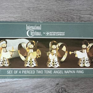 May include: A set of four pierced two-tone angel napkin rings in a box. The angels are gold-coloured with white details and hold instruments. The box reads "International Christmas" and "SET OF 4 PIERCED TWO TONE ANGEL NAPKIN RING".