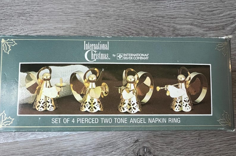 May include: A set of four pierced two-tone angel napkin rings in a box. The angels are gold-coloured with white details and hold instruments. The box reads "International Christmas" and "SET OF 4 PIERCED TWO TONE ANGEL NAPKIN RING".