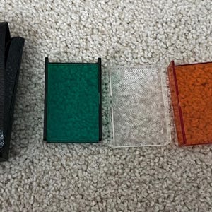 Peut inclure: Un étui en cuir noir avec une fermeture à bouton-pression, à côté de quatre filtres de caméra colorés : vert, transparent, orange et rouge. Les filtres sont rectangulaires avec des cadres noirs. L'étui et les filtres sont sur une surface de couleur neutre.
